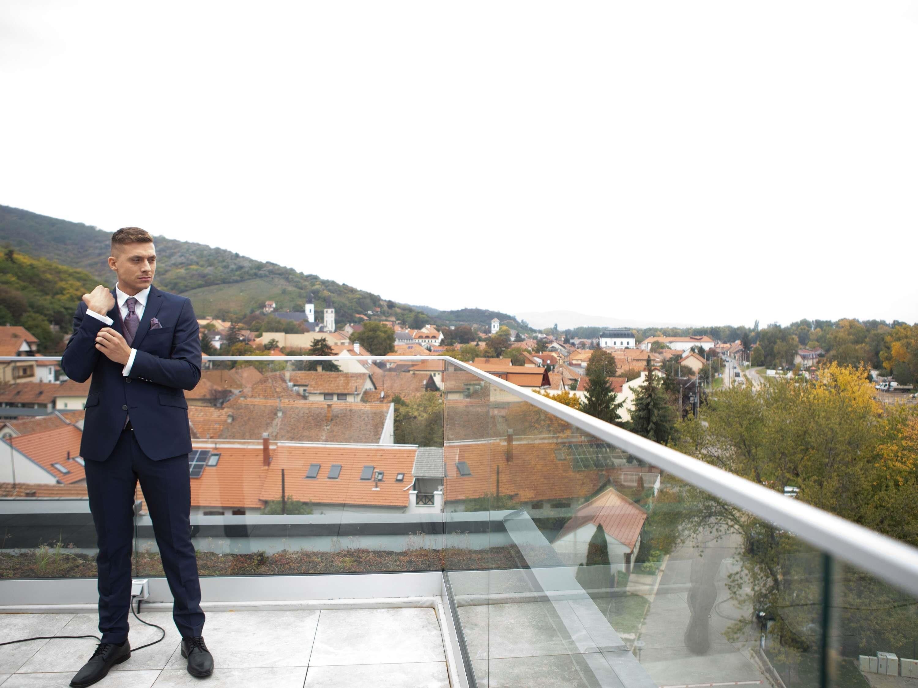 Un hombre con traje de pie en un balcón, con vistas a un paisaje urbano.