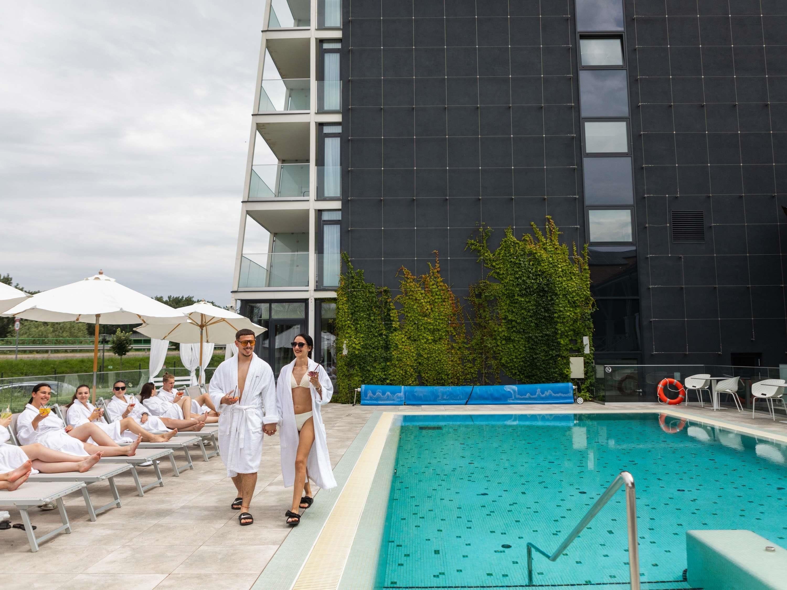 Un área de piscina con sillones de descanso y sombrillas, una piscina y un edificio moderno con balcones.