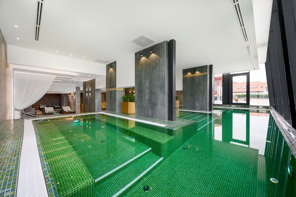 A modern indoor swimming pool with a green tiled floor, large pillars, and a view of the outside. - Mercure Tokaj Center (Tokaj)
