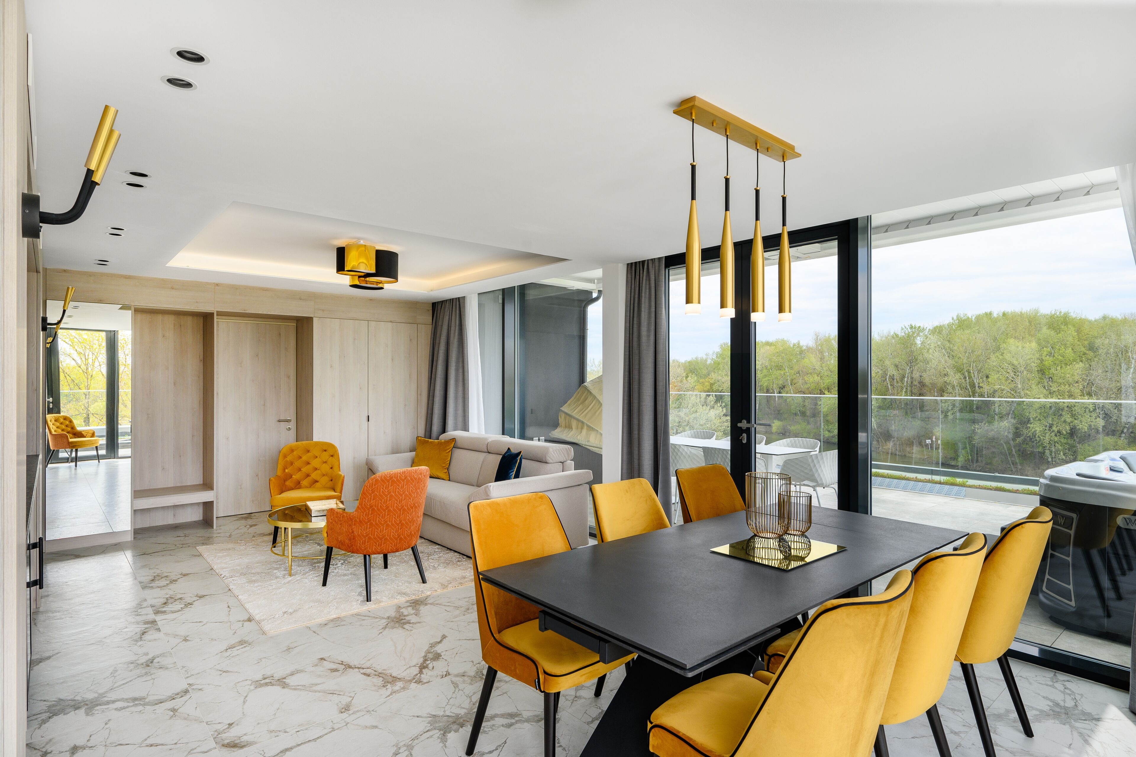 Et moderne spiseområde med et sort bord og gule stole, en sofa med orange og hvide puder, samt udsigt til grøn vegetation udenfor.