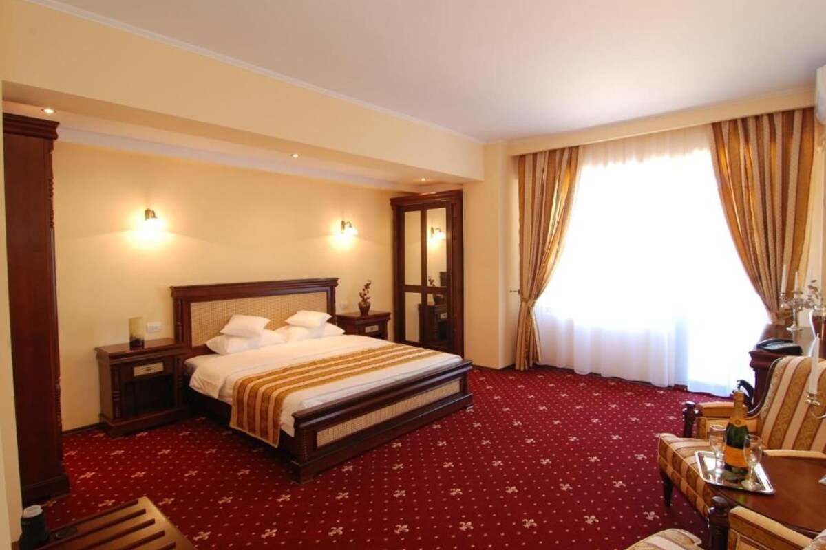 A hotel room with a large bed, a wooden headboard, a nightstand, a bedside table, a chair, a table with a bottle and glasses, and a window with curtains.