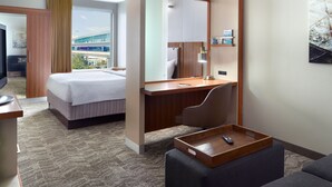 A hotel room with a bed, a desk, a chair, a tray table, and a large window offering a view of a building and greenery.