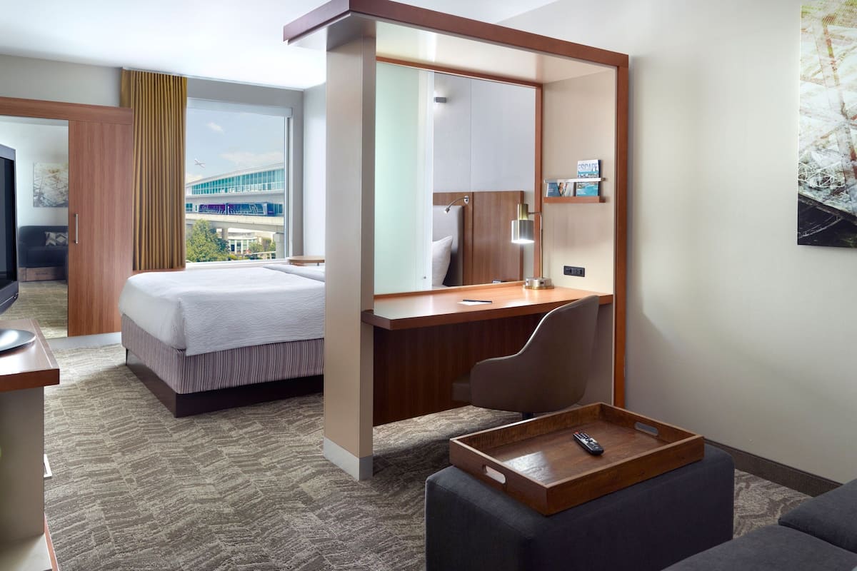 A hotel room with a bed, a desk, a chair, a tray table, and a large window offering a view of a building and greenery.