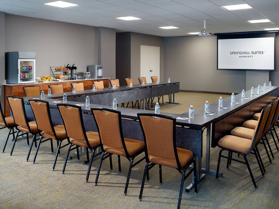 A conference room with a long table, chairs, a refrigerator with beverages, and a screen displaying 'SPRINGHILL SUITES® MARRIOTT'.