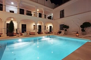 A swimming pool with a clear blue water surface, surrounded by white columns and arches. There are potted plants and outdoor seating areas.