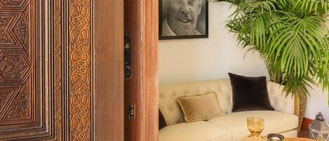 Una habitación con una puerta de madera, un sofá color crema, una mesa redonda de madera, una planta grande en maceta y un cuadro en la pared.