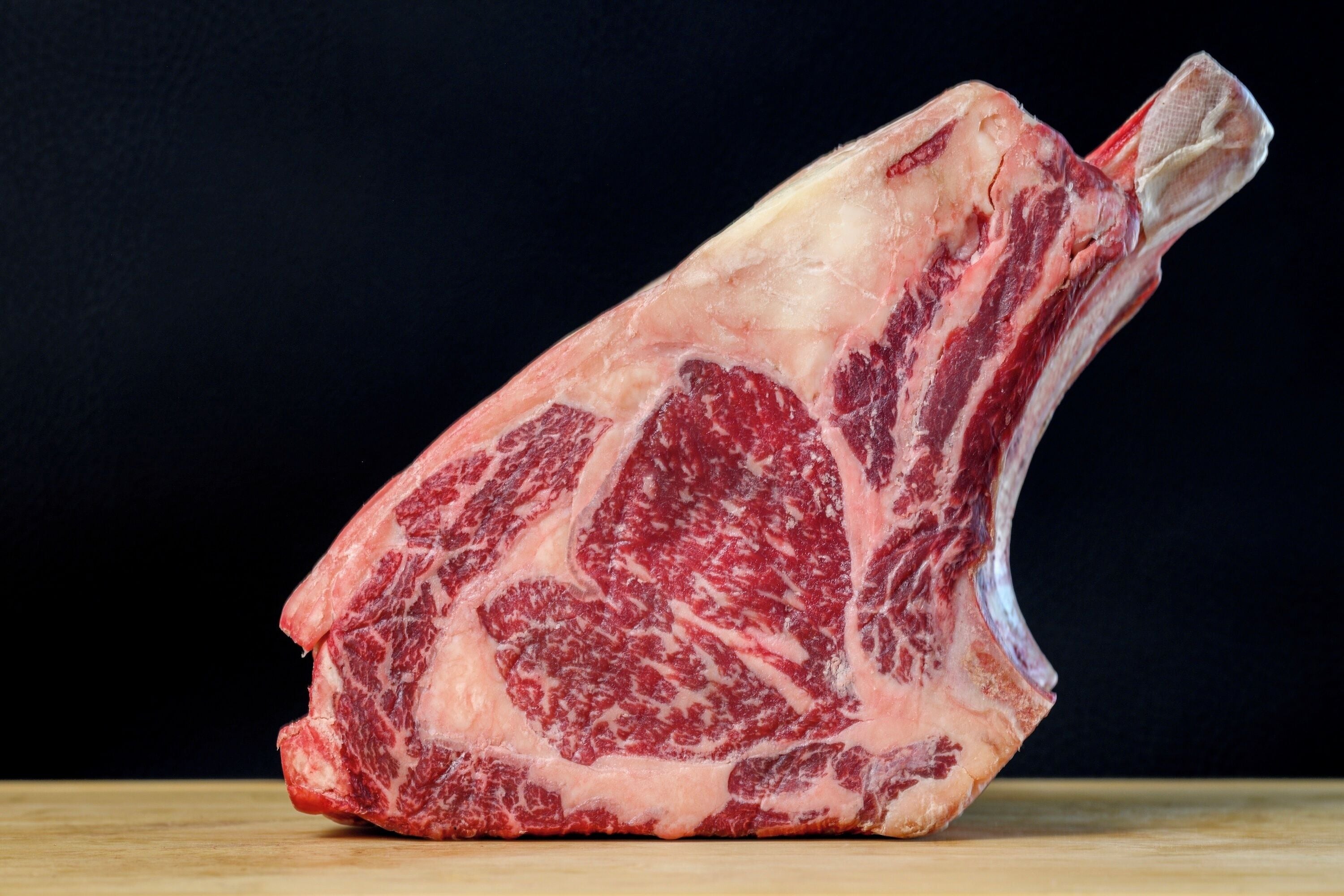A large, raw piece of meat, likely a rib cut, on a wooden surface against a dark background.