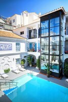A modern building with a glass facade, a swimming pool, and a courtyard with lounge chairs and potted plants.