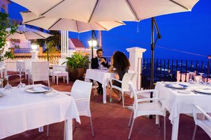 An outdoor dining area with white tables and chairs, large umbrellas, and potted plants.