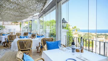 A restaurant with outdoor seating, white tables, blue cushions, and a view of the sea.