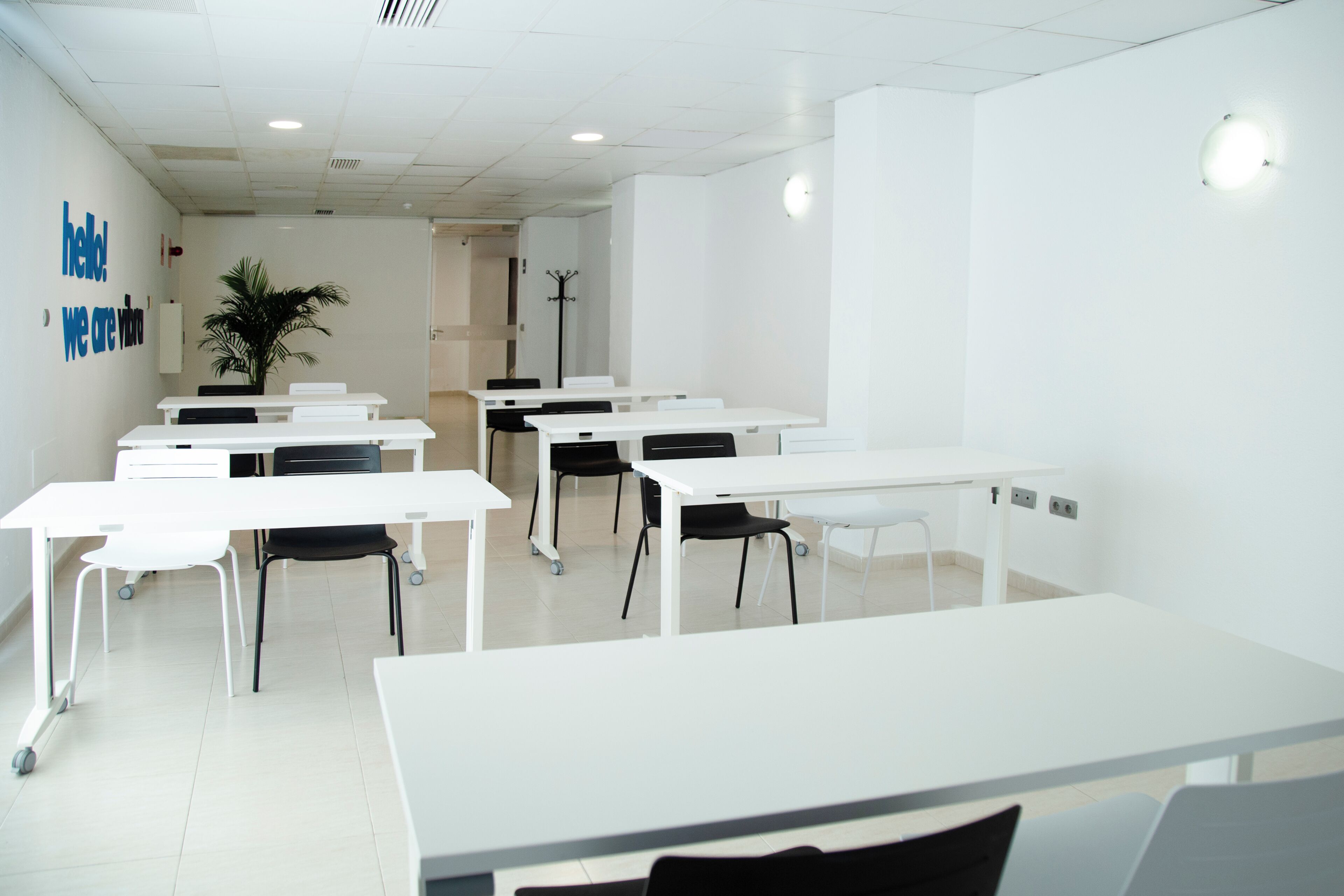 Een vergaderzaal met witte tafels en stoelen, een potplant en een muur met de tekst 'hello, we are here'.