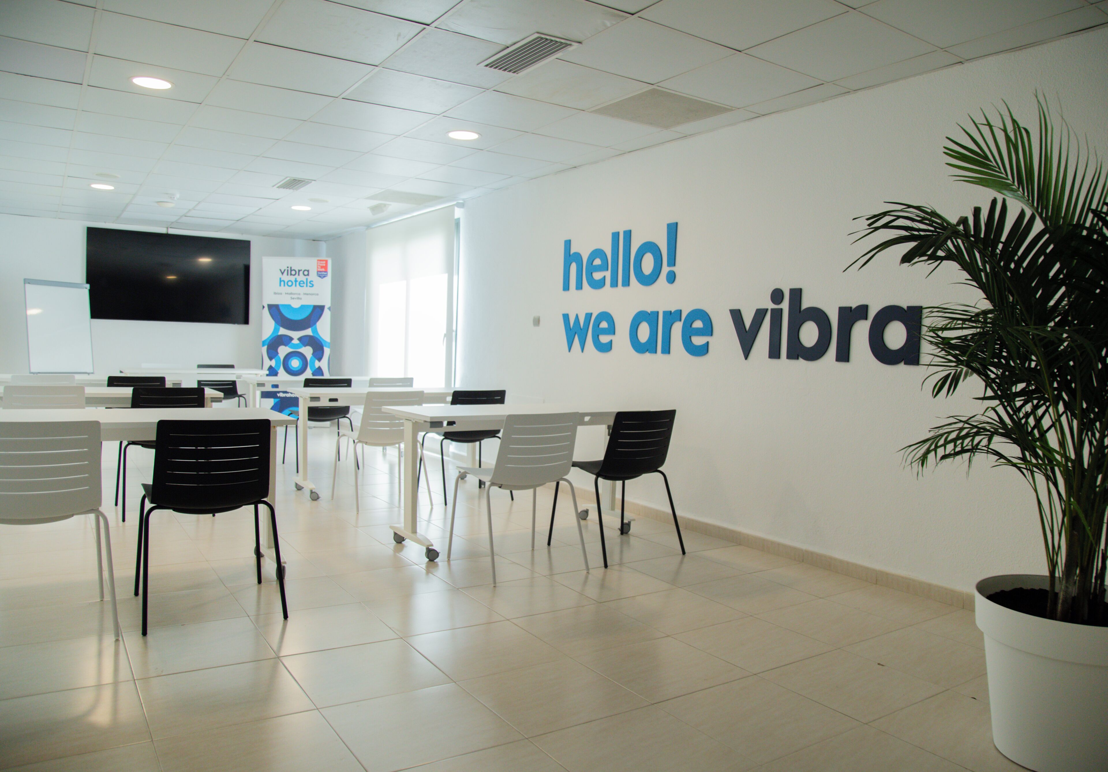 A modern hotel lobby with a reception desk, chairs, a large wall sign, and a potted plant.