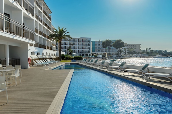 Una piscina con sillas de descanso y terraza de madera, rodeada de edificios modernos y palmeras.