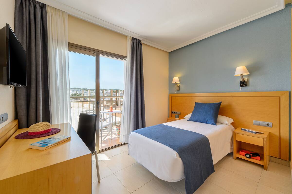 Una habitación de hotel con una cama, un escritorio con un sombrero y un libro, un televisor, una mesita de noche con una l ámpara y vistas a la ciudad a través de la ventana.