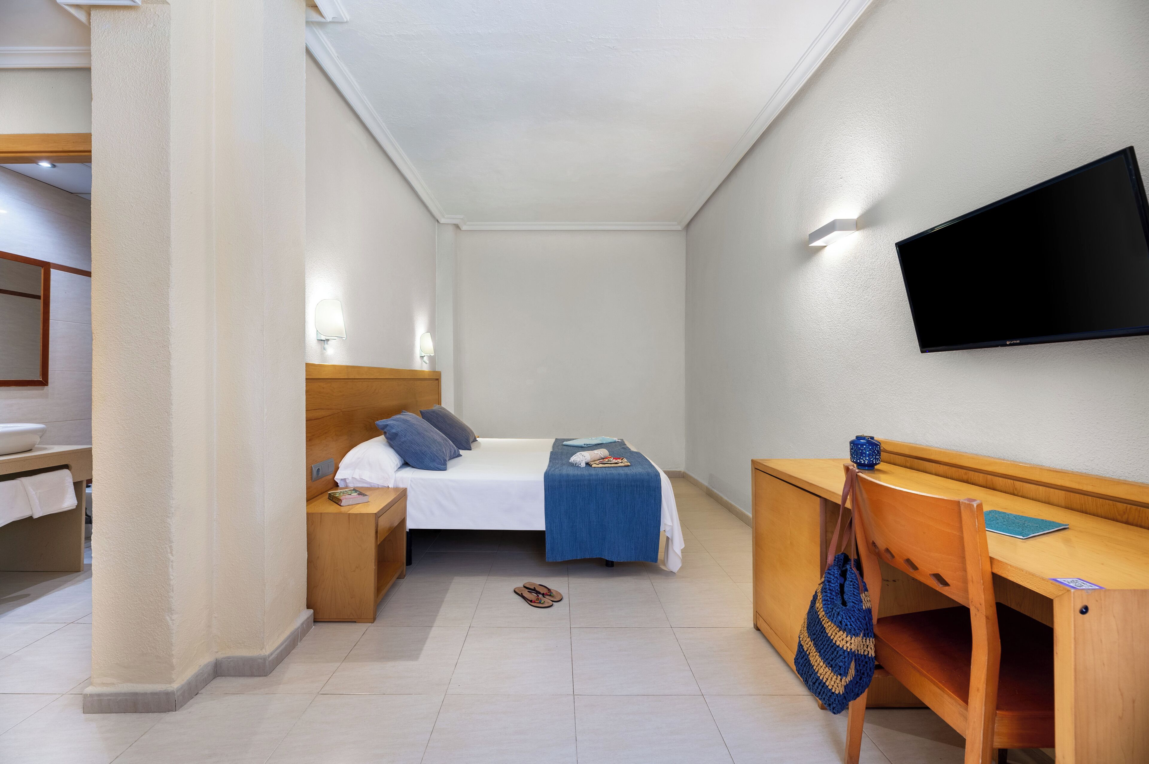A hotel room with a bed, bedside table, lamp, TV, desk, chair, and a wooden wardrobe.
