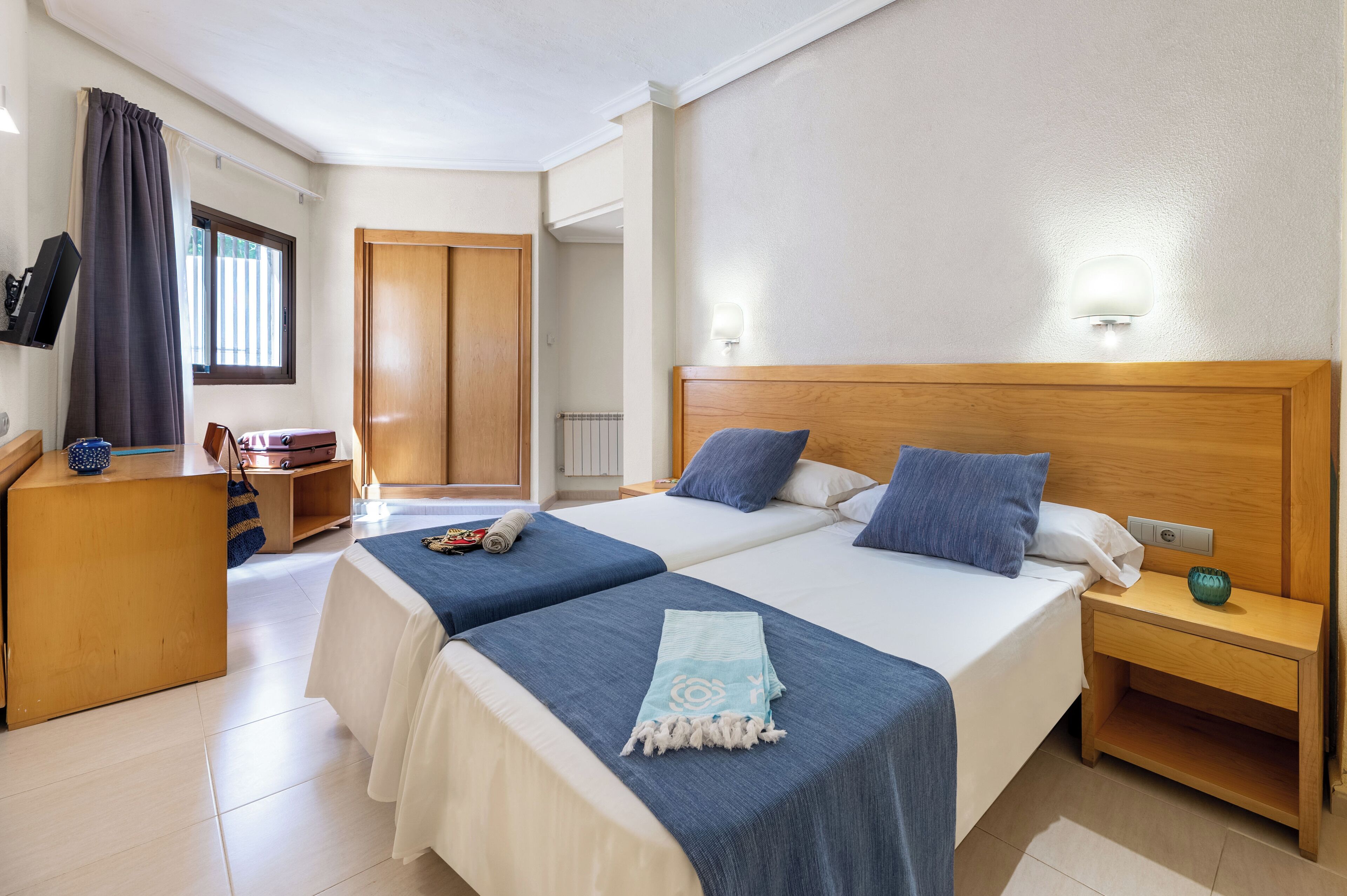A hotel room with two beds, a wooden nightstand, a desk, and a TV.