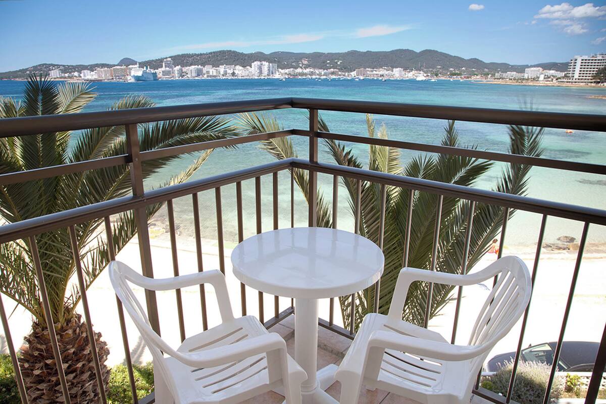 Un balcón con una mesa blanca y dos sillas, con vista a una playa de aguas azul turquesa y palmeras.