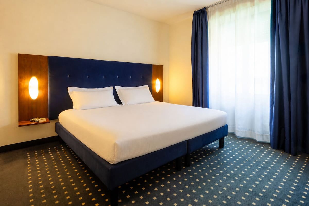 A hotel room with a large bed, two bedside tables, a headboard, and a window with curtains.