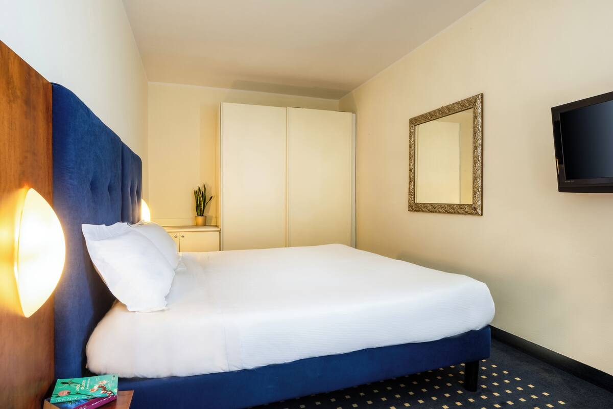 A hotel room with a bed, a bedside lamp, a mirror, a TV, and a small table with a book.