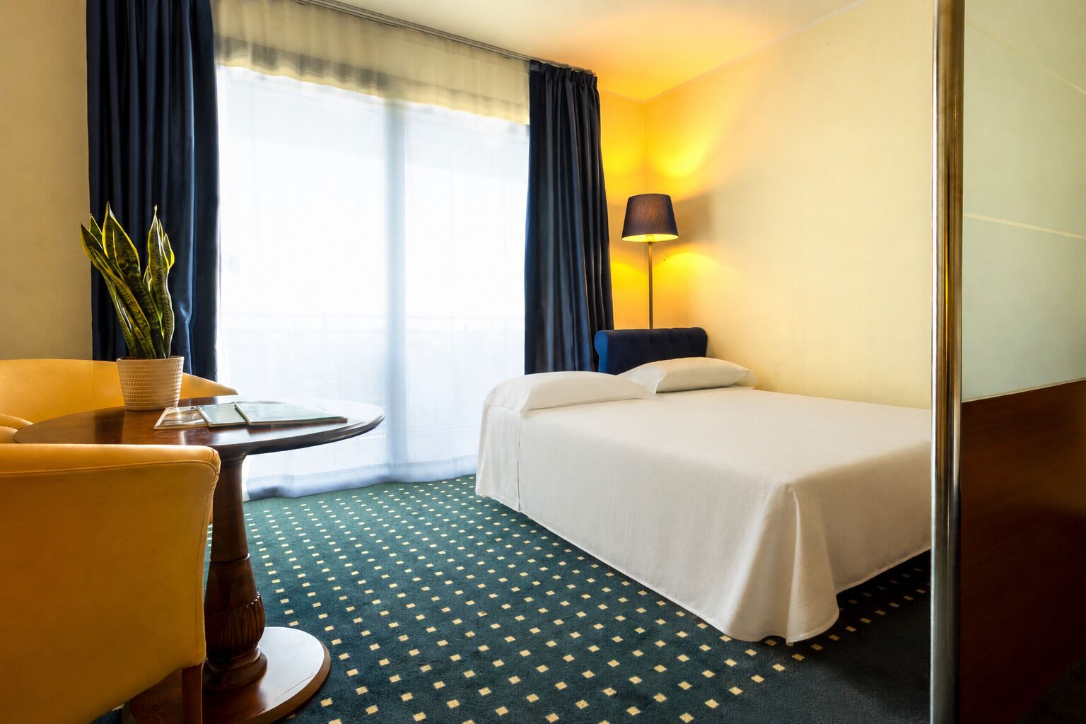 A hotel room with a bed, a desk with a chair, a lamp, and a potted plant.