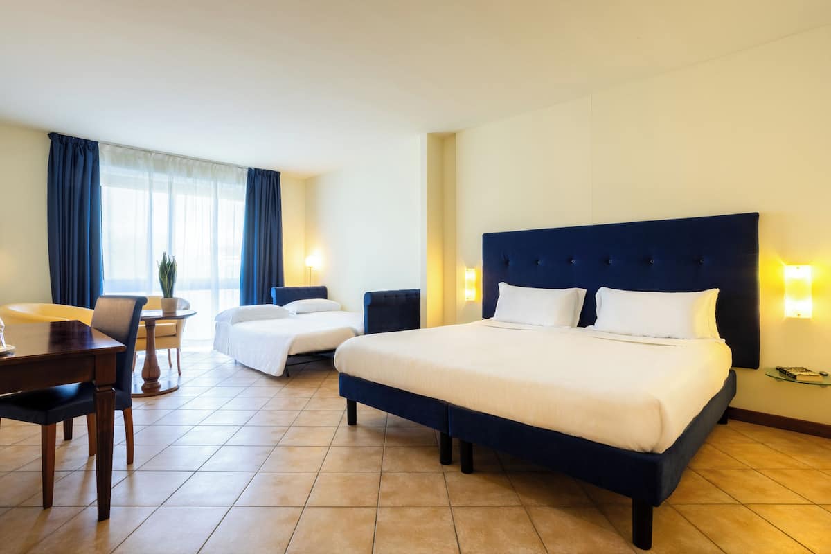 A hotel room with a large bed, two smaller beds, a dining table, chairs, a potted plant, and a window with curtains.