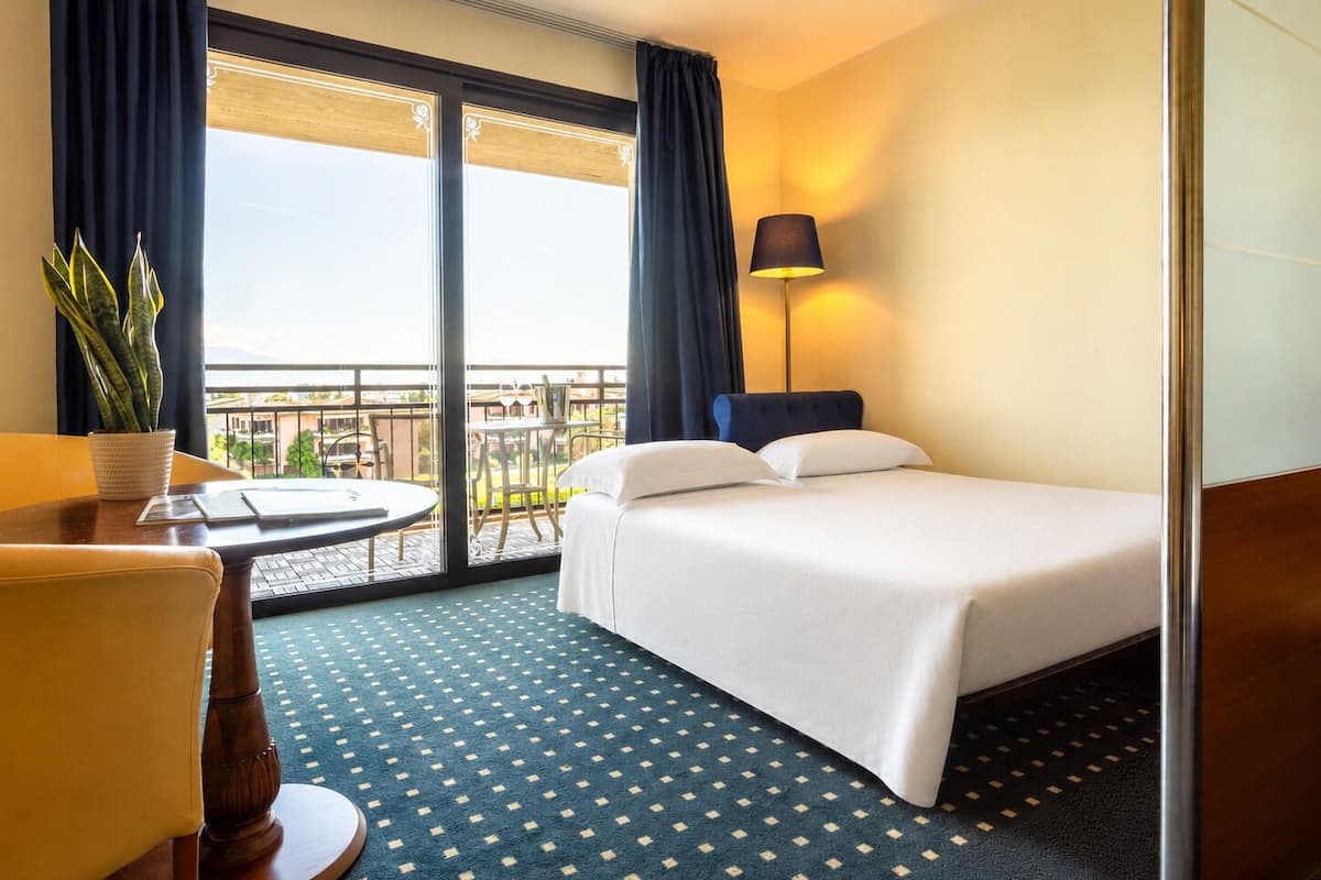 A hotel room with a bed, a desk, a chair, a potted plant, a table, and a balcony with a view.