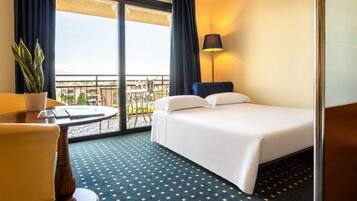 A hotel room with a bed, a desk, a chair, a potted plant, a table, and a balcony with a view.