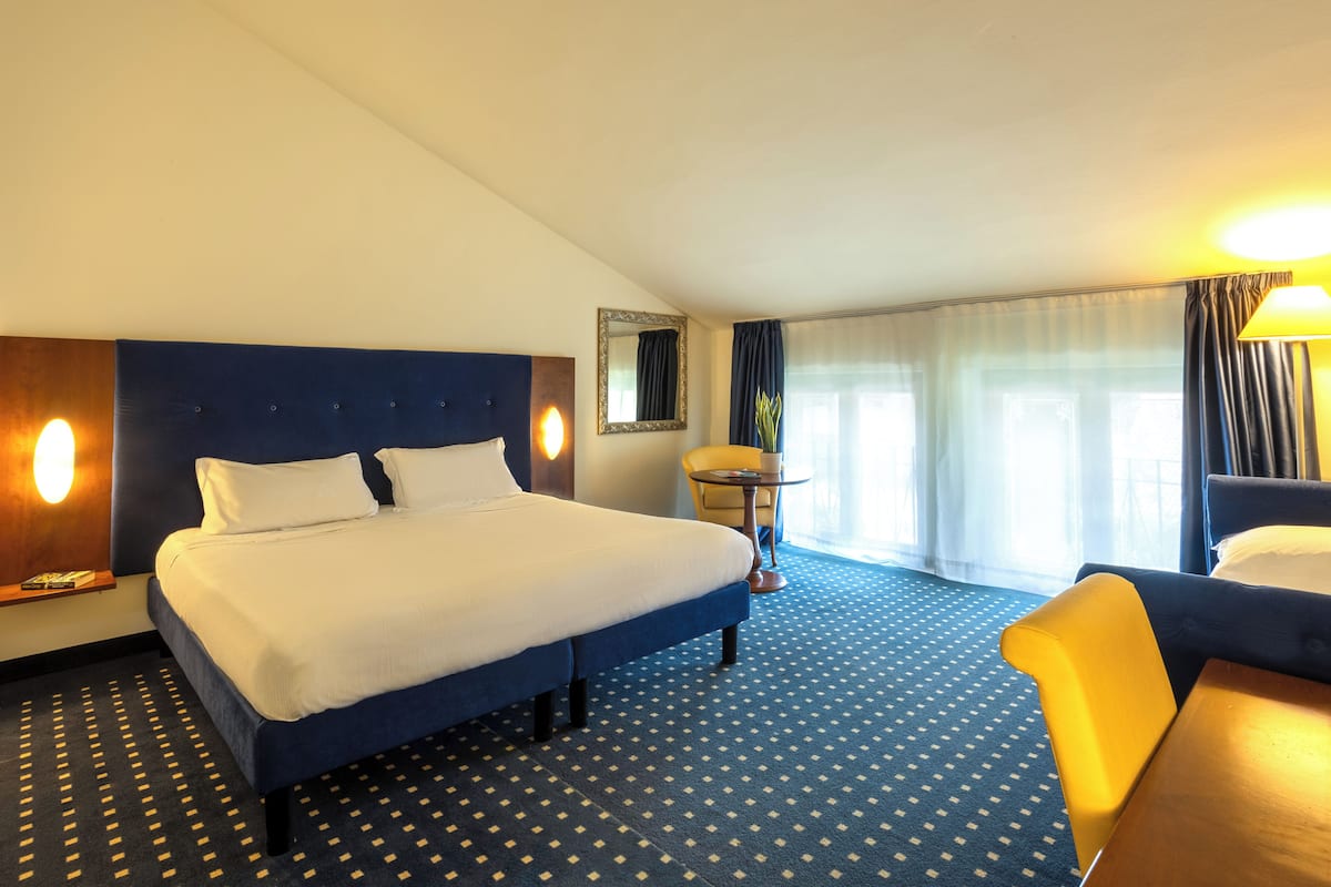 A hotel room with a large bed, a small table, a chair, and a window with curtains.