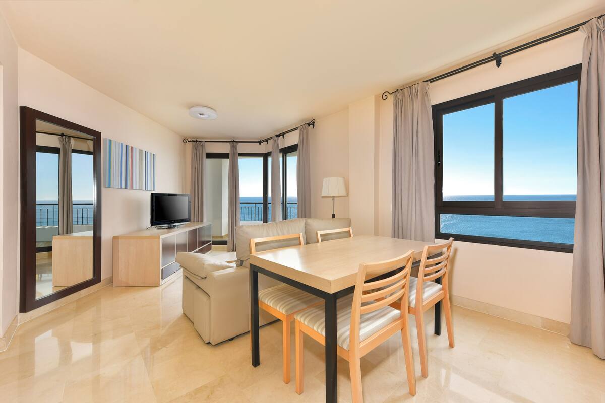 Une chambre d’hôtel moderne dotée d’un coin repas, d’une télévision et offrant une vue sur la mer.
