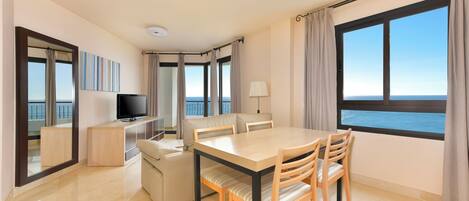 A modern hotel room with a dining area, a TV, and a view of the sea.