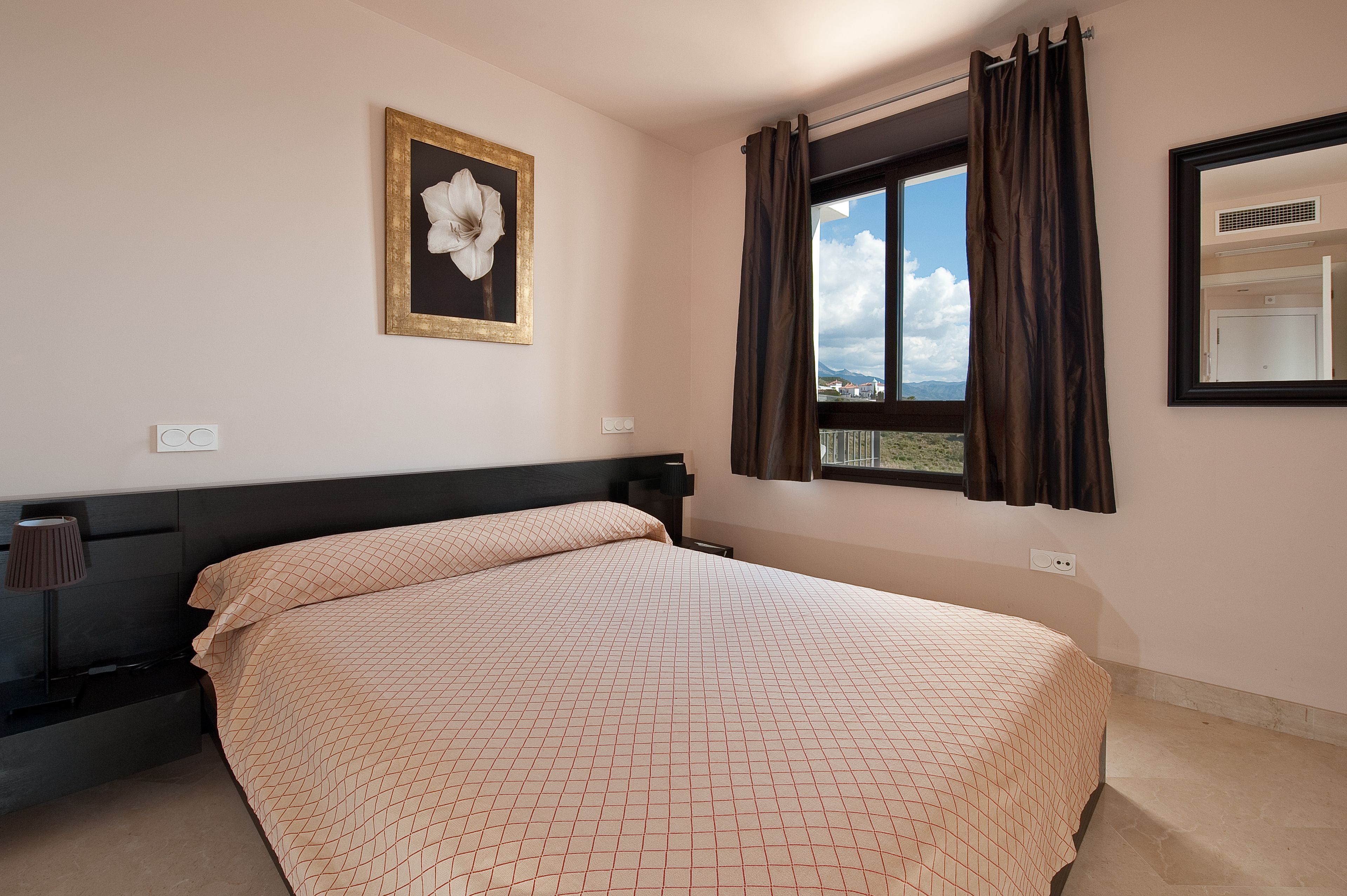 A bedroom with a large bed, a nightstand, a lamp, a framed picture, a window with curtains, and a view of the outside.