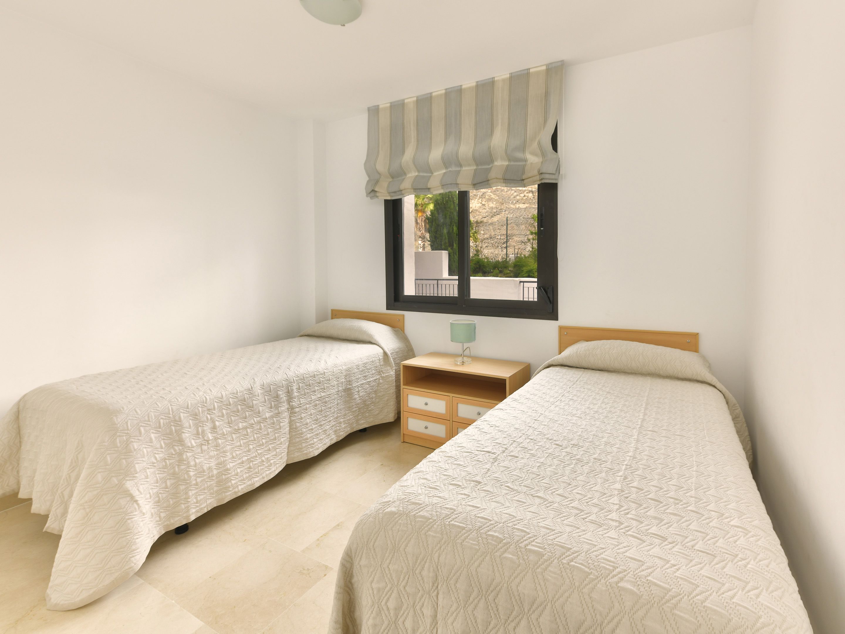 Une chambre avec deux lits simples, une table de chevet, une lampe et une fenêtre donnant sur l’extérieur.