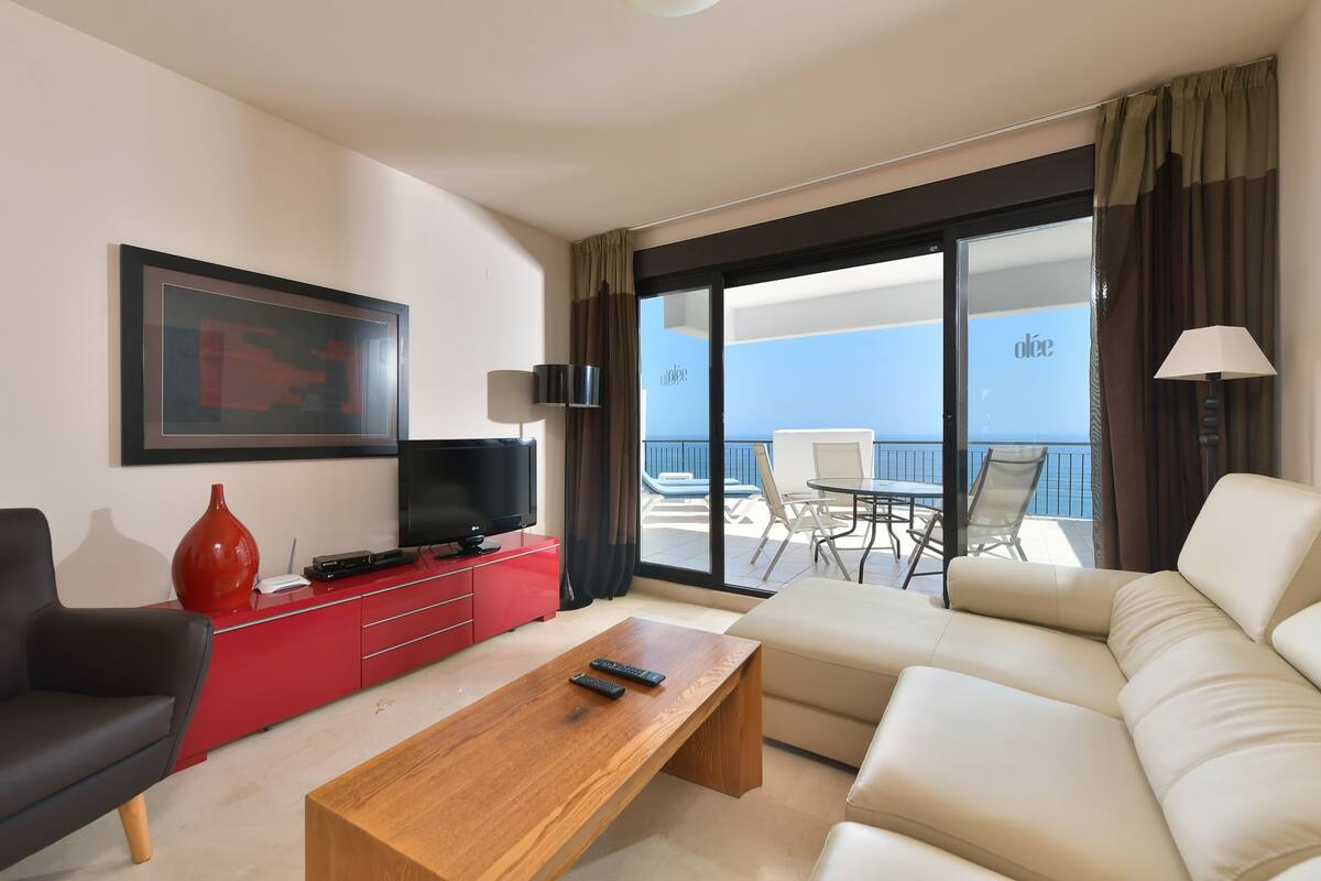 Une chambre d’hôtel moderne avec un grand canapé, une table basse en bois, un meuble de rangement rouge, une télévision et un balcon équipé d’une table et de chaises.