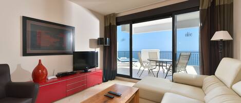 A modern hotel room with a large sofa, a wooden coffee table, a red entertainment unit, a TV, and a balcony with a table and chairs.