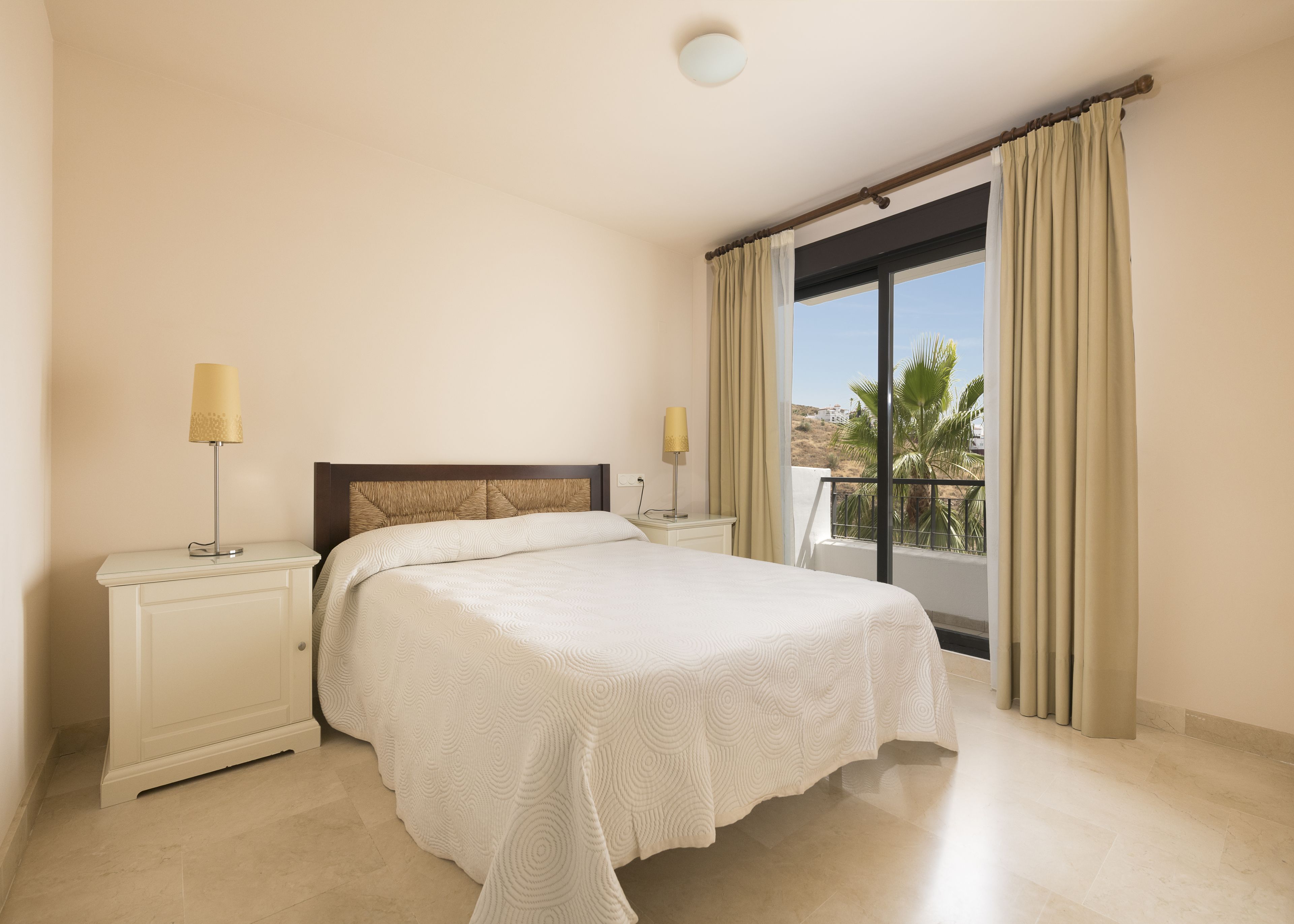 A bedroom with a bed, two bedside tables, a window with curtains, and a view of palm trees.