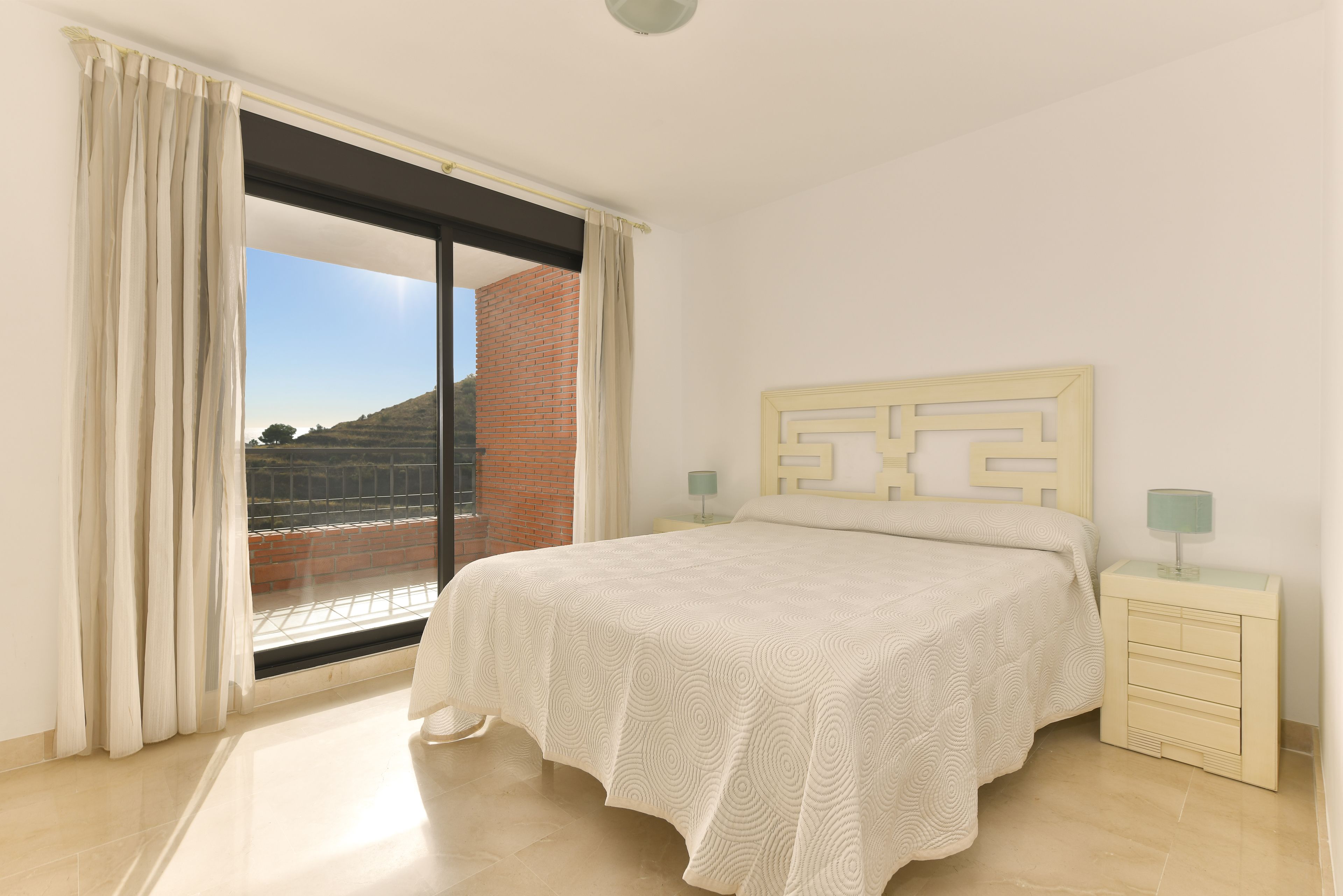 A bedroom with a large window, a bed with a patterned bedspread, a nightstand with a lamp, and a wall with a geometric design.
