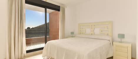 A bedroom with a large window, a bed with a patterned bedspread, a nightstand with a lamp, and a wall with a geometric design.