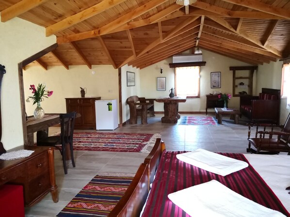 A spacious attic room with a high wooden ceiling, a dining area with a table and chairs, a bed with a striped bedspread, and a refrigerator.