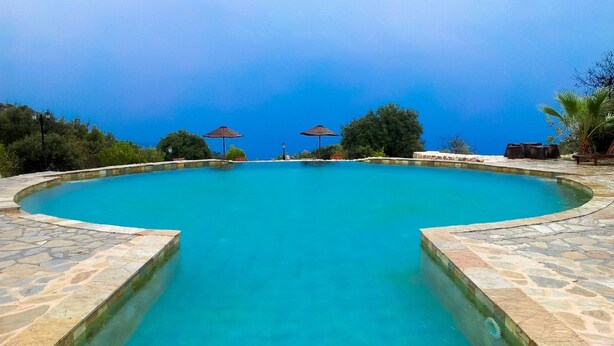 Une grande piscine d’un bleu éclatant, bordée de pierres, entourée d’arbres et de parasols.
