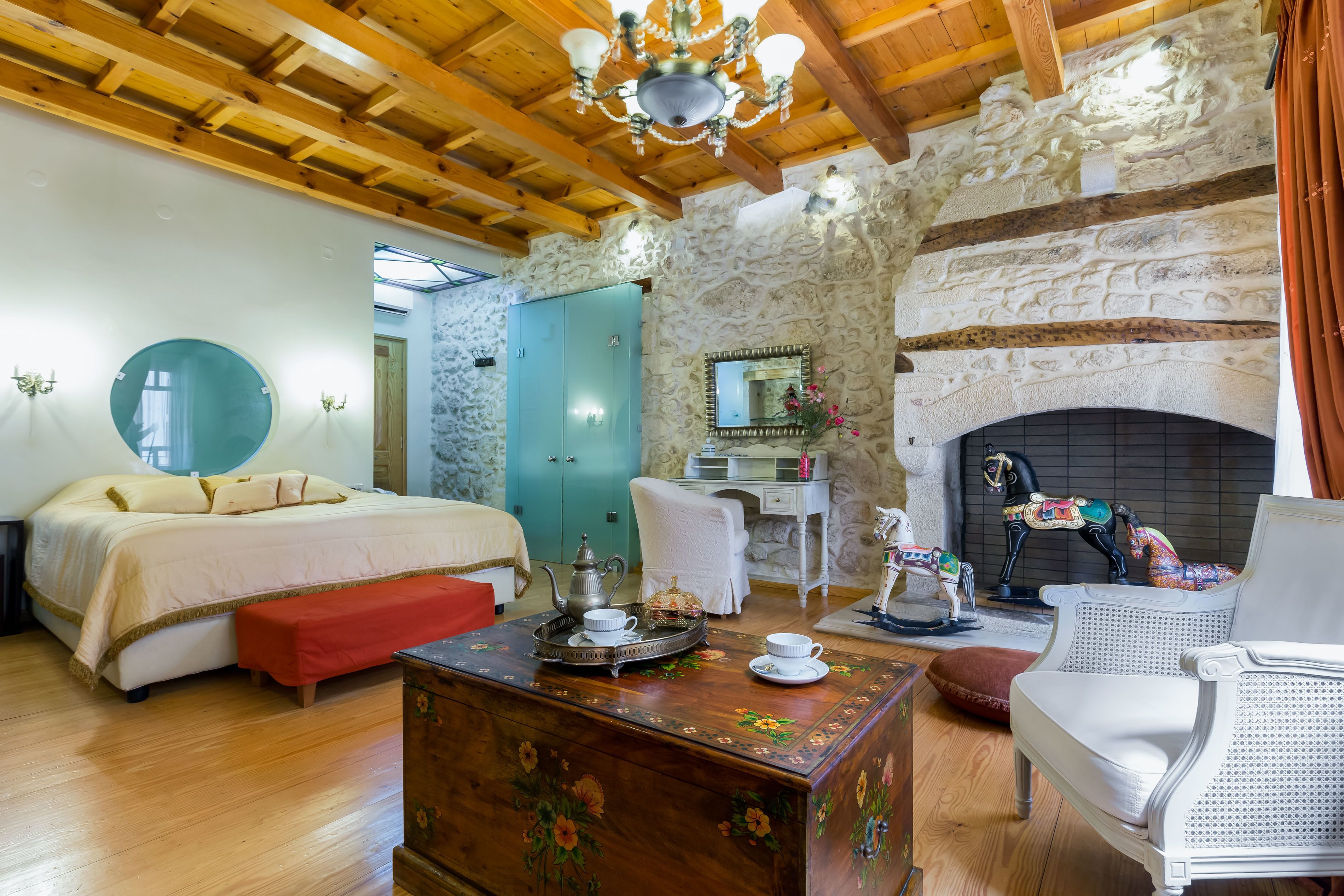 A spacious room with a large bed, a wooden ceiling, a stone wall, a fireplace, and a dining area with a table set for two.