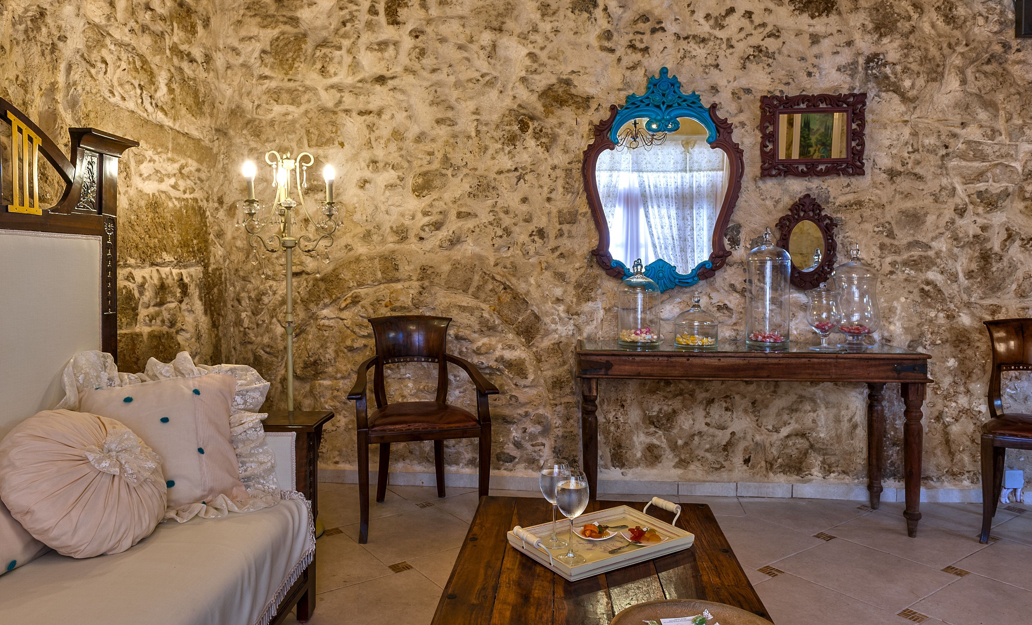 A room with a stone wall, a wooden chair, a sofa with decorative pillows, a wooden table with a tray of food and glasses, and a mirror with ornate frames.