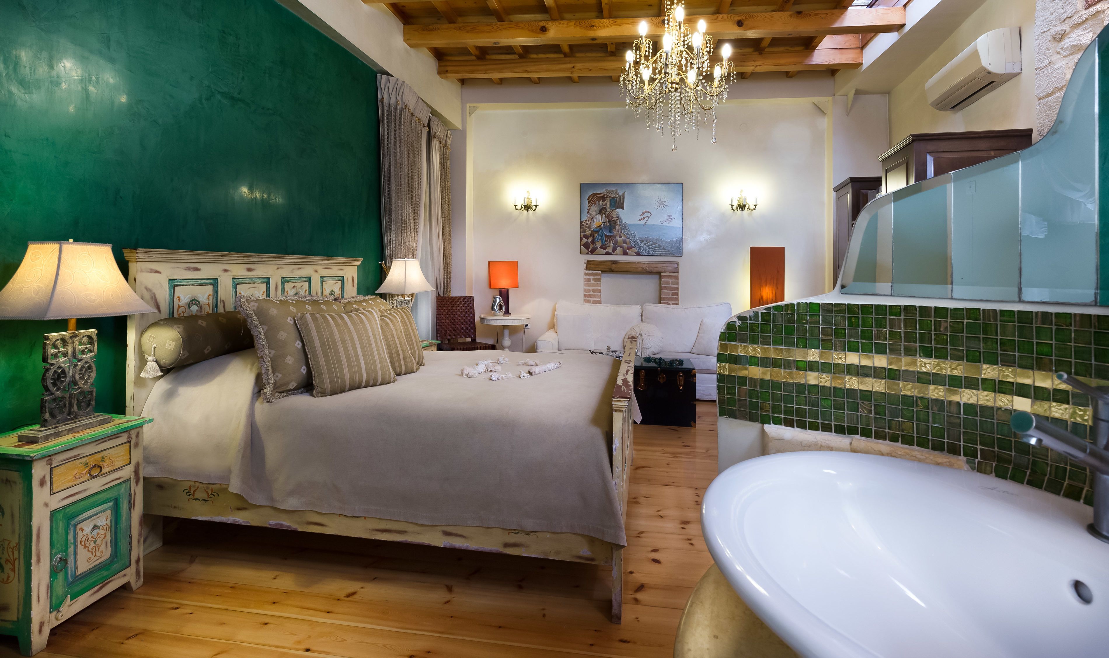 A bedroom with a bed, bedside tables, a chandelier, a painting, and a bathroom with a sink and mirror.