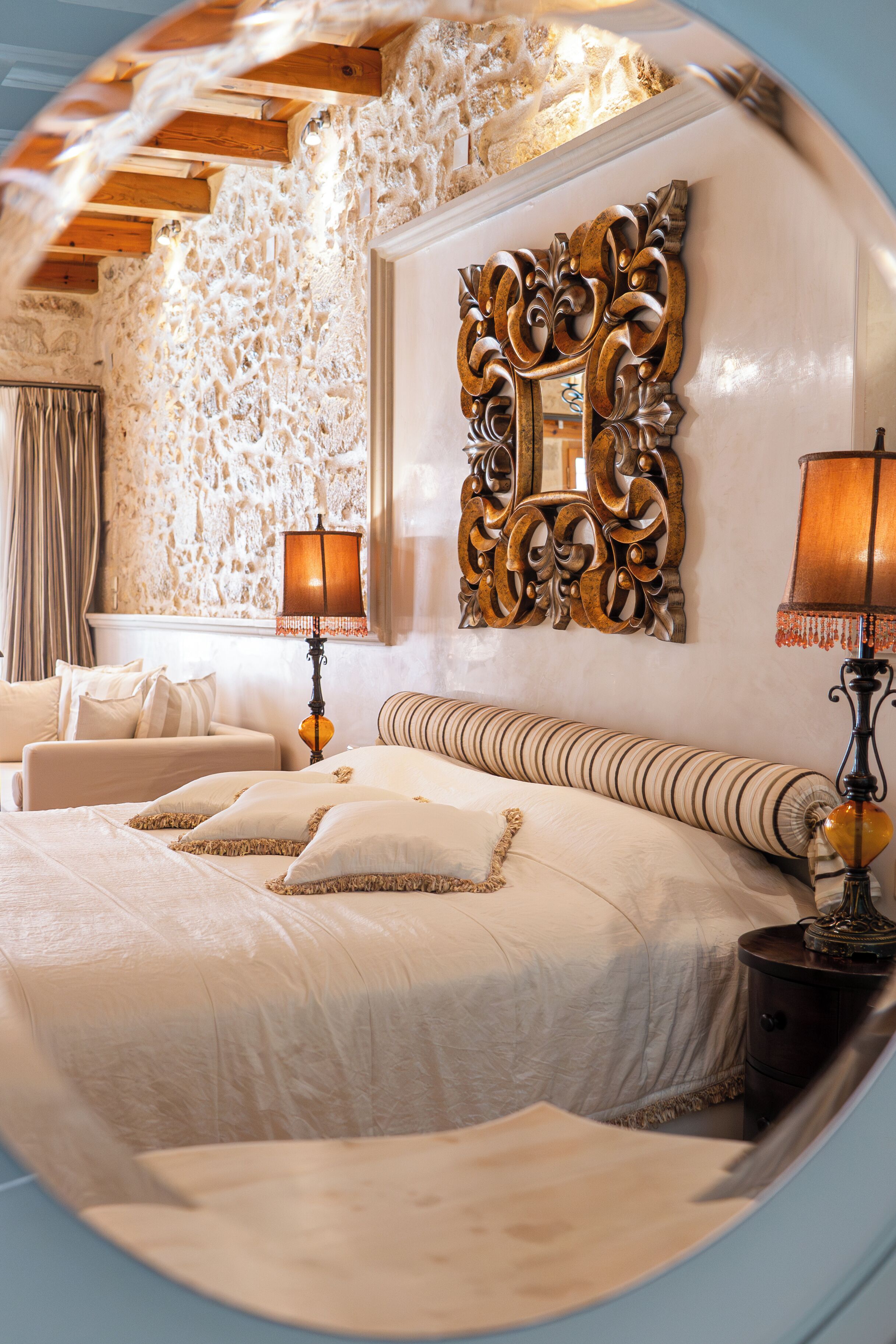 Un dormitorio con pared de piedra, un espejo grande y ornamentado, dos lámparas de noche y una cama con muchos almohadones.