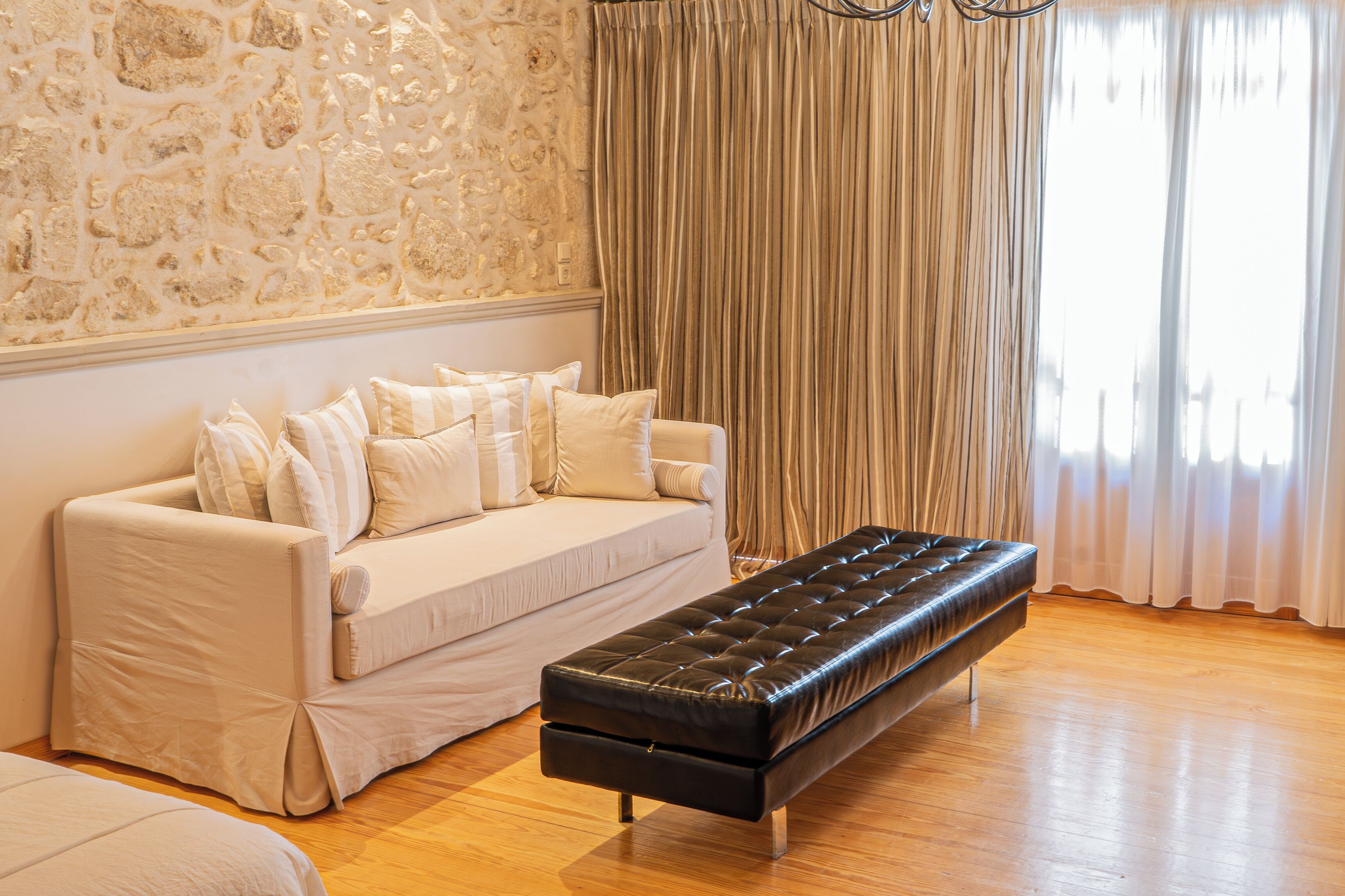 Una habitación con sofá, banco, pared de piedra y ventana con cortinas.