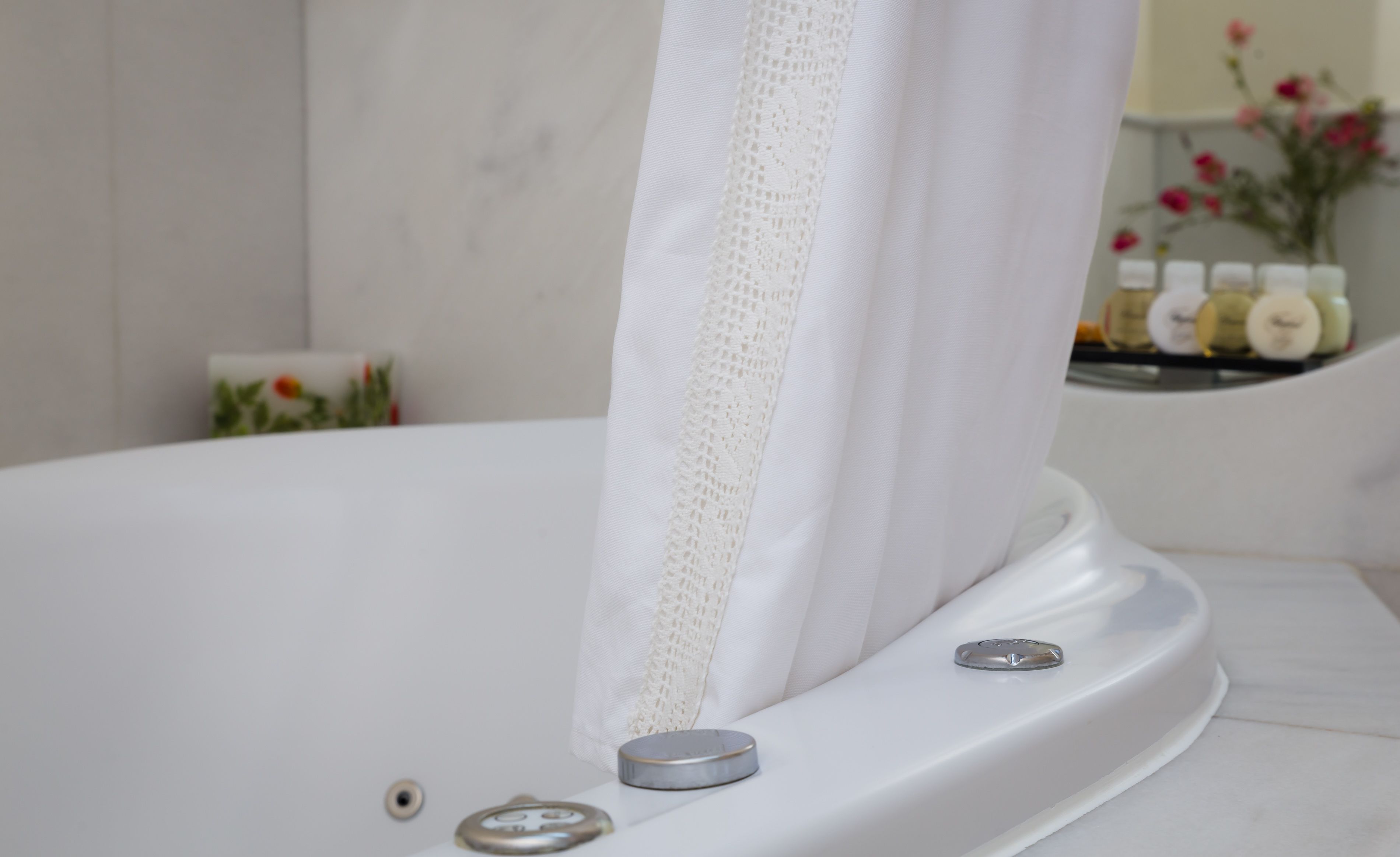 A white bathtub with a lace curtain, a control knob, and a small bottle of shampoo on the edge.