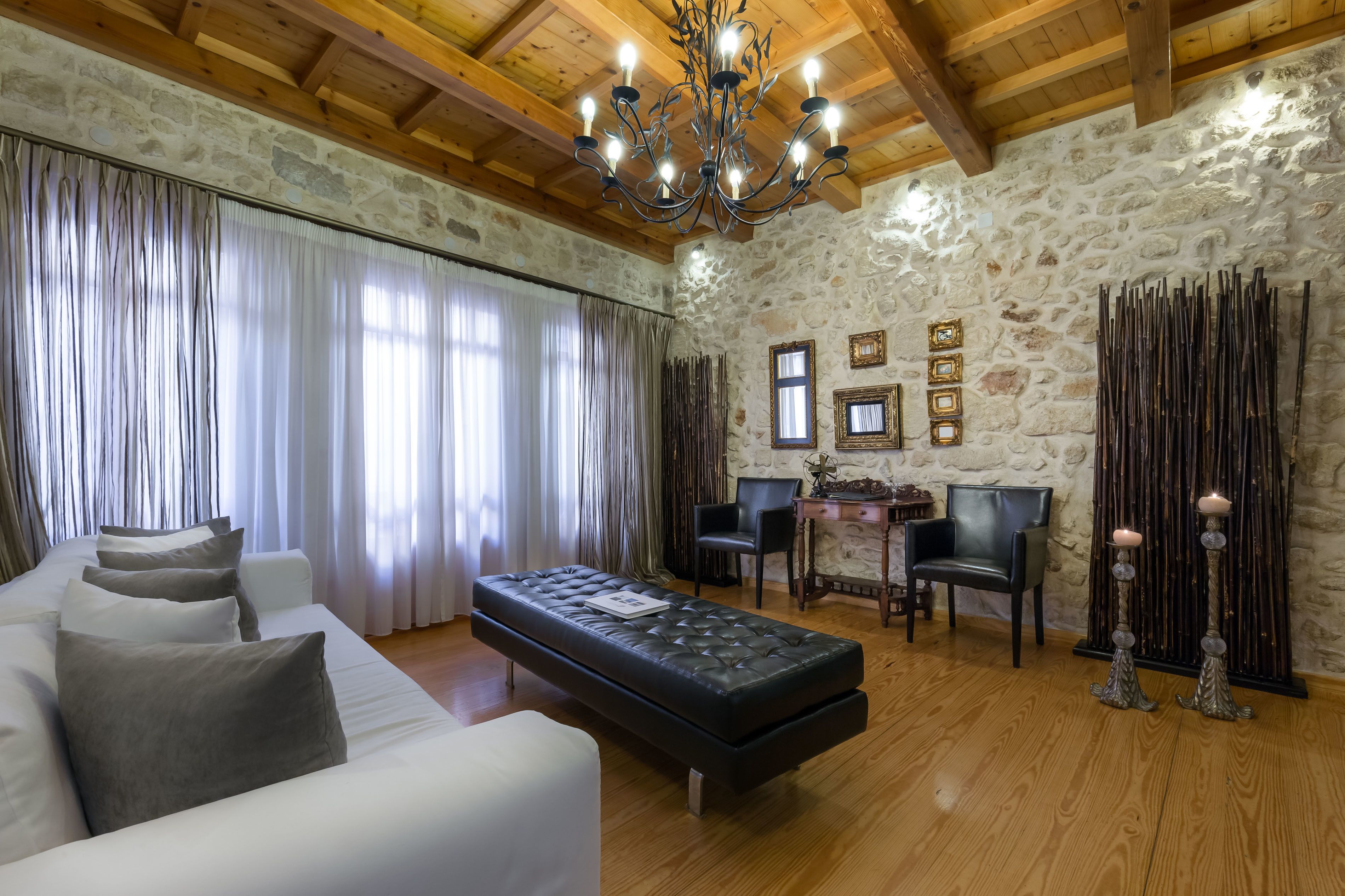 Una habitación con pared de piedra, techo de madera, un otomano negro, sofá blanco y una araña.