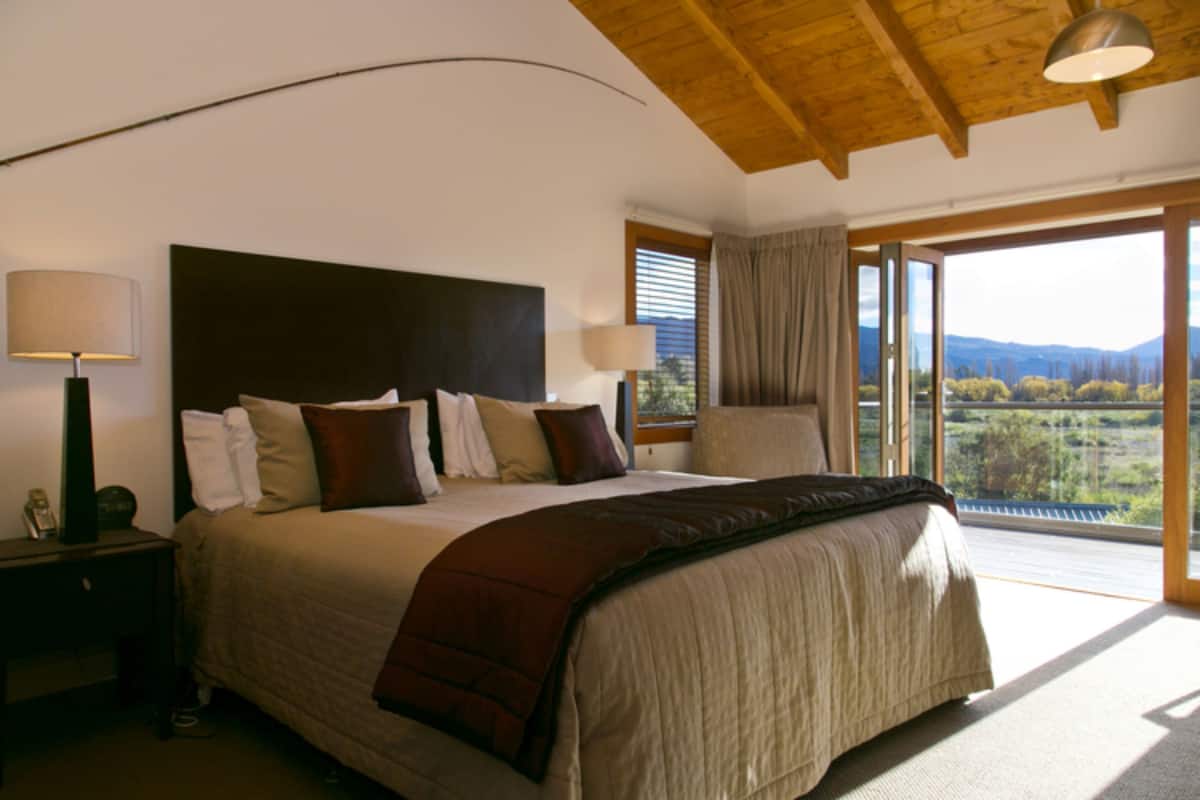 A bedroom with a large bed, a nightstand, a lamp, and a view of the outdoors through a large window.