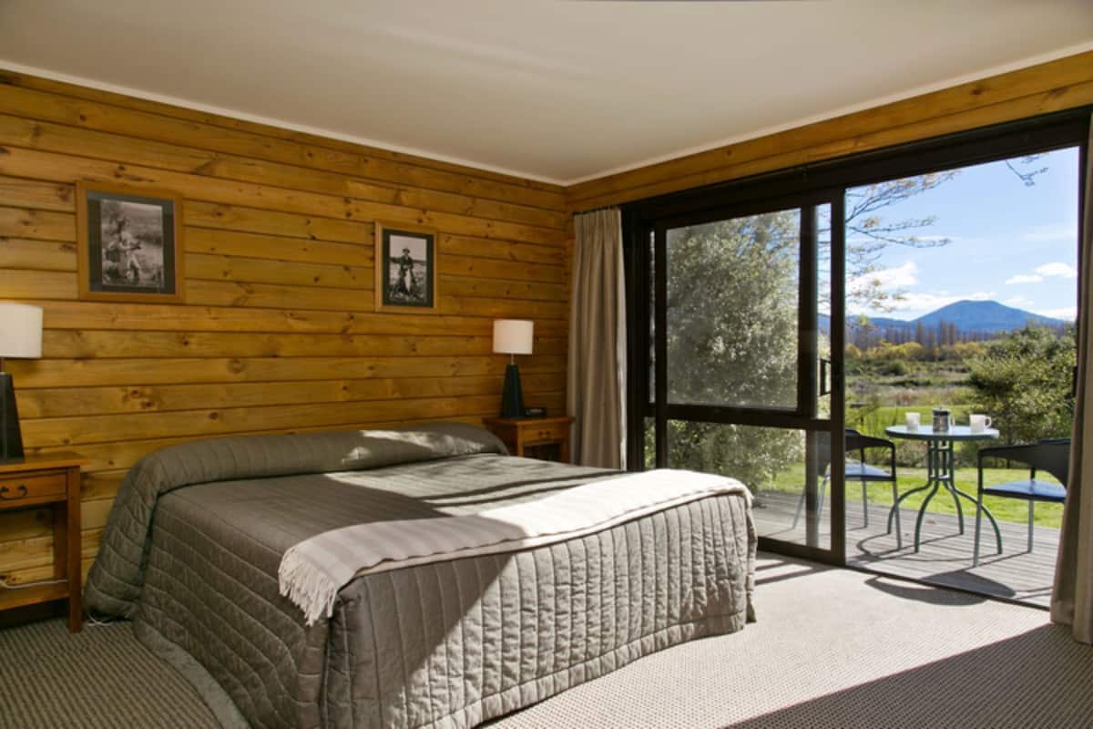 A bedroom with a large bed, wooden walls, a window, and a view of a garden.