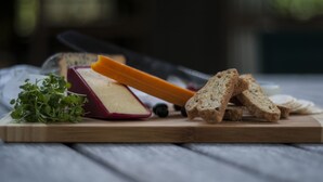A wooden cutting board with a wedge of cheese, slices of bread, and a small bunch of greens.
