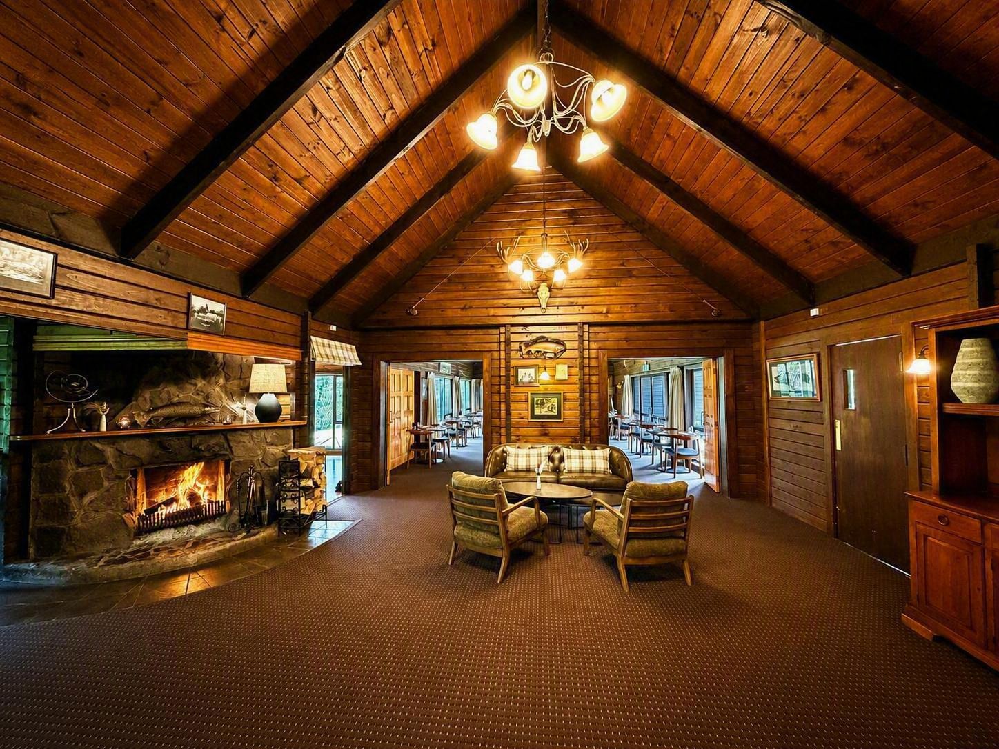 A log cabin interior with a stone fireplace, wooden ceiling beams, a dining area with a round table and chairs, and a chandelier.
